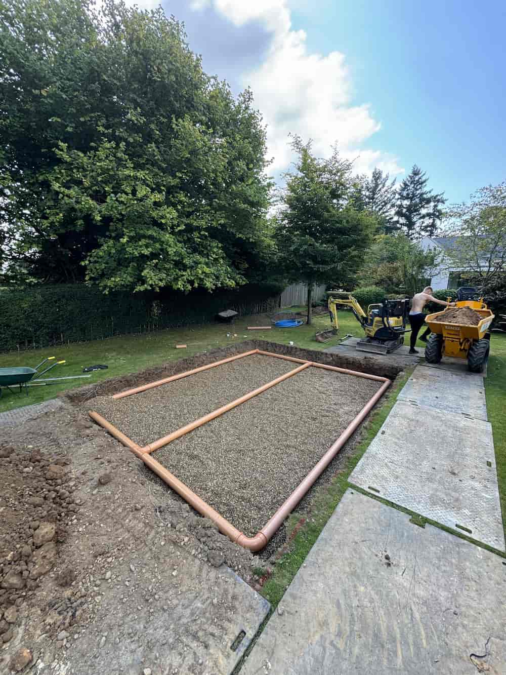 A rectangular gravel foundation with exposed piping is being built in a grassy yard. Construction equipment, including a small digger and dumper, is nearby, and trees border the work site under a blue sky.
