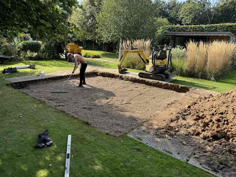 A person spreads gravel with a rake on a rectangular patch of ground, next to a small yellow excavator in a grassy garden surrounded by tall grasses and trees.