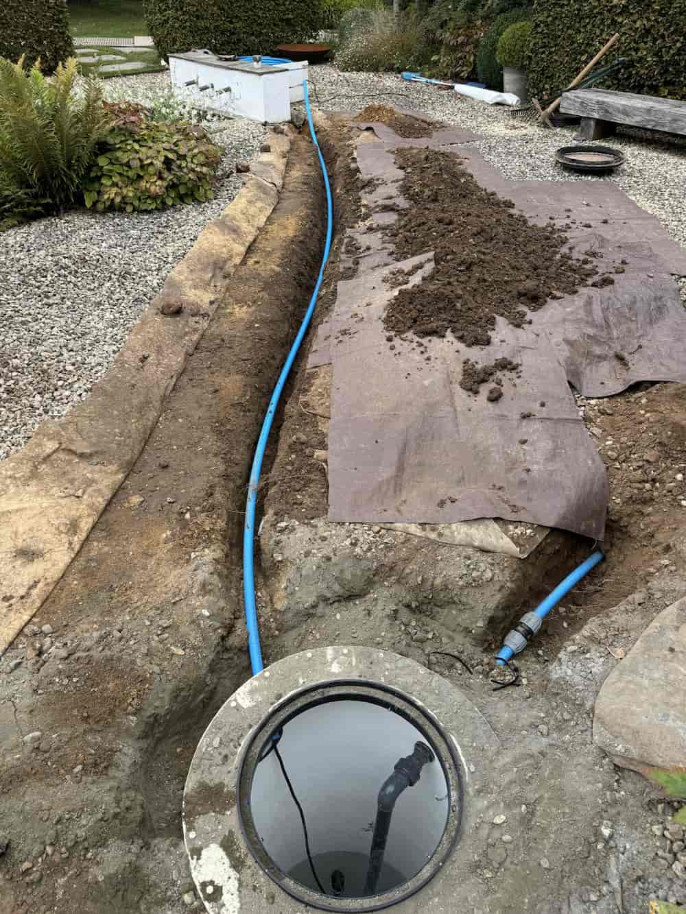 A blue pipe runs through a trench in a gravel and soil garden, leading to a circular water meter or access cover. Soil is piled on one side, and protective sheets are laid out along the trench. Shrubs and plants are nearby.