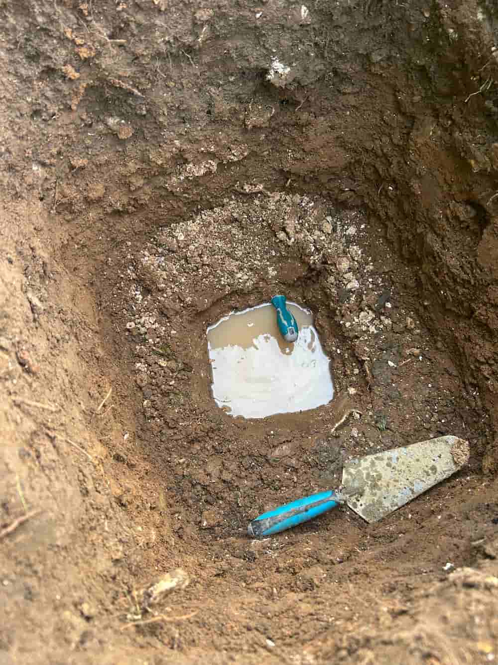 A muddy hole in the ground partially filled with water, containing a blue-handled small tool and another blue-handled trowel resting on the soil nearby.