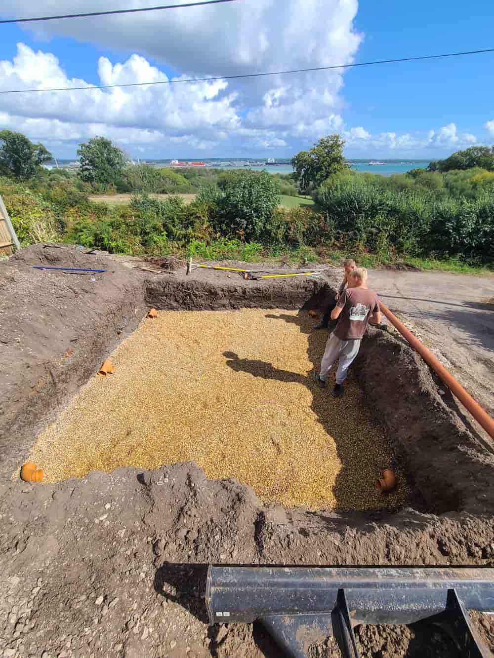 A person stands in a large rectangular pit lined with gravel, surrounded by dirt mounds, with trees and greenery in the background under a partly cloudy sky.