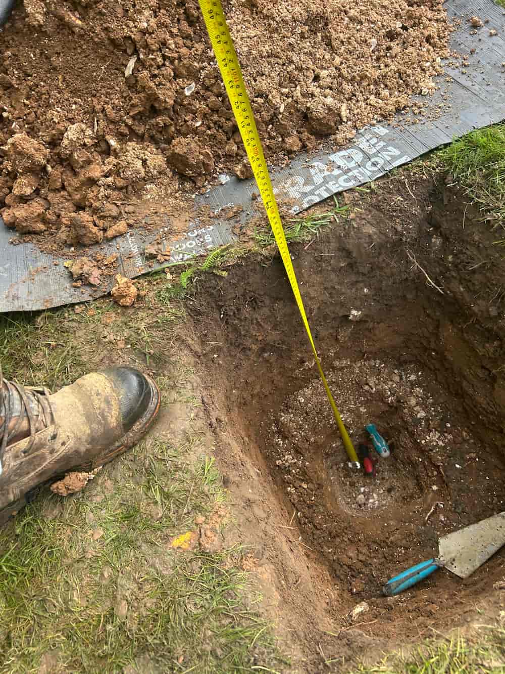 A person in work boots stands beside a dug hole in the ground, with a yellow measuring tape extended into the hole. Soil, tools, and a mat are visible around the site.