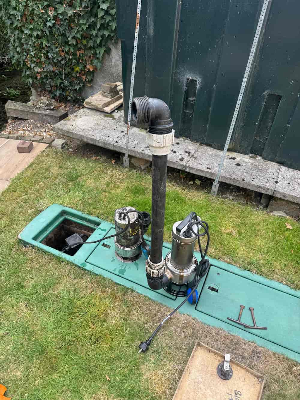 Two submersible pumps are placed on an open green underground tank cover in a grassy yard, with hoses and cables attached; tools and bricks are nearby, and a black shed is in the background.