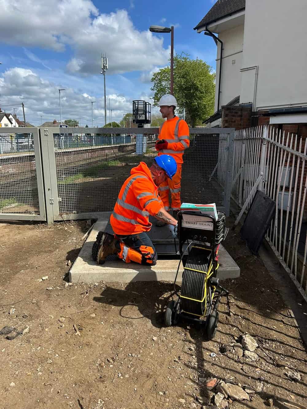 Two construction workers in orange safety gear and helmets work outdoors near a metal fence, one kneeling by an open ground hatch with tools, the other standing and observing. Equipment and buildings are visible in the background.