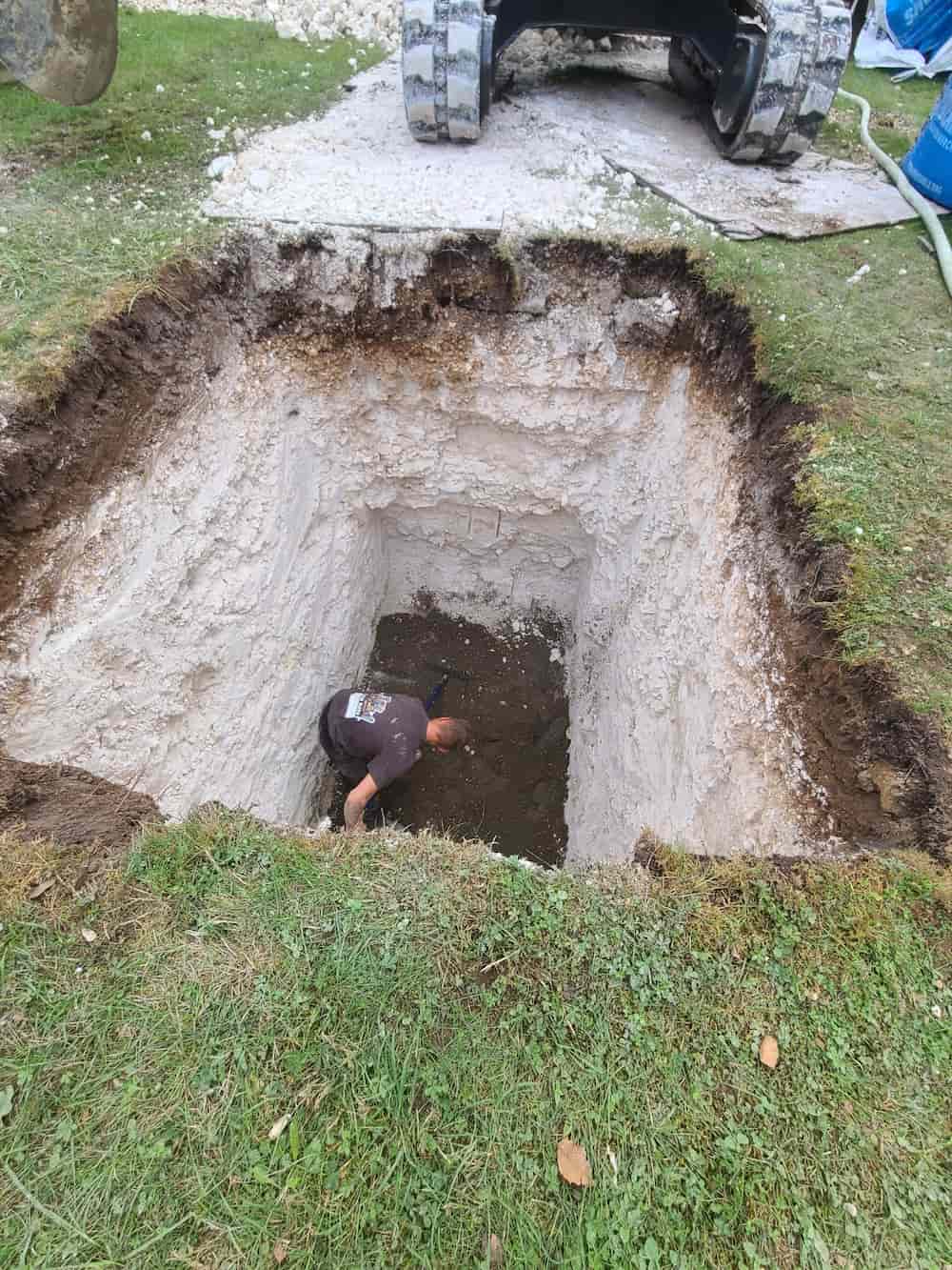 A person is working at the bottom of a large, deep rectangular pit dug in grassy ground, with an excavator partially visible at the top edge of the hole.