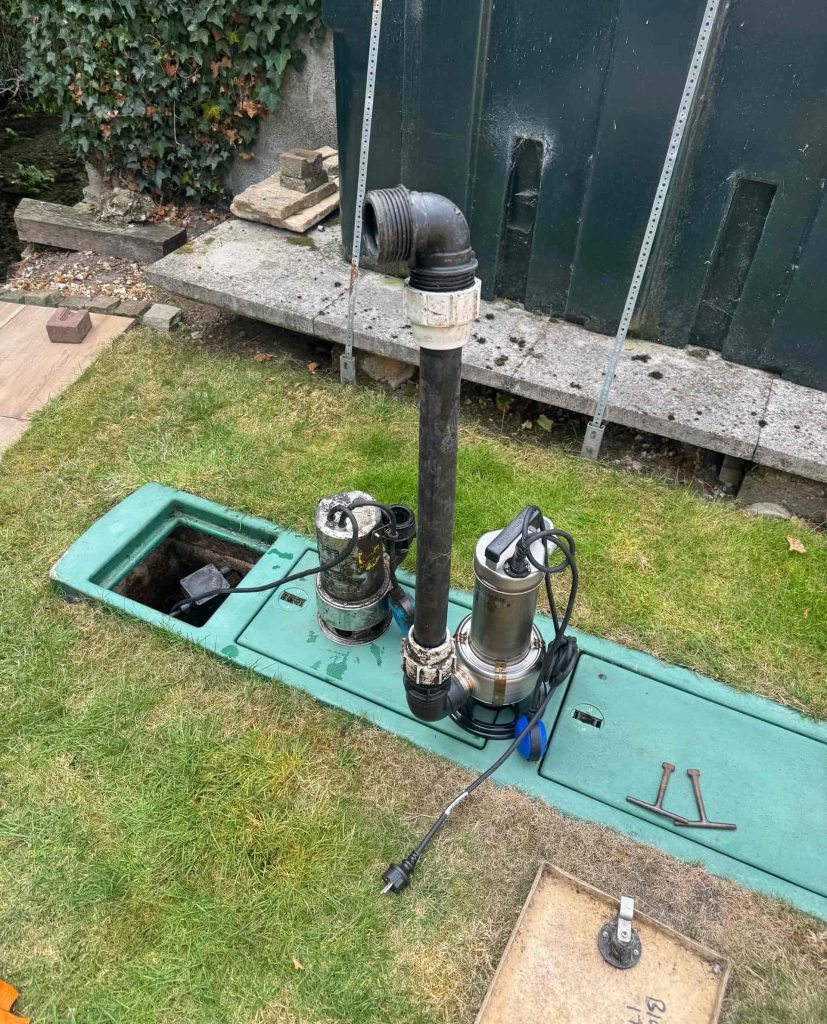 Two submersible water pumps sit on a green underground tank cover in a grassy yard, with cables and pipes attached. An open hatch and tools are visible nearby.