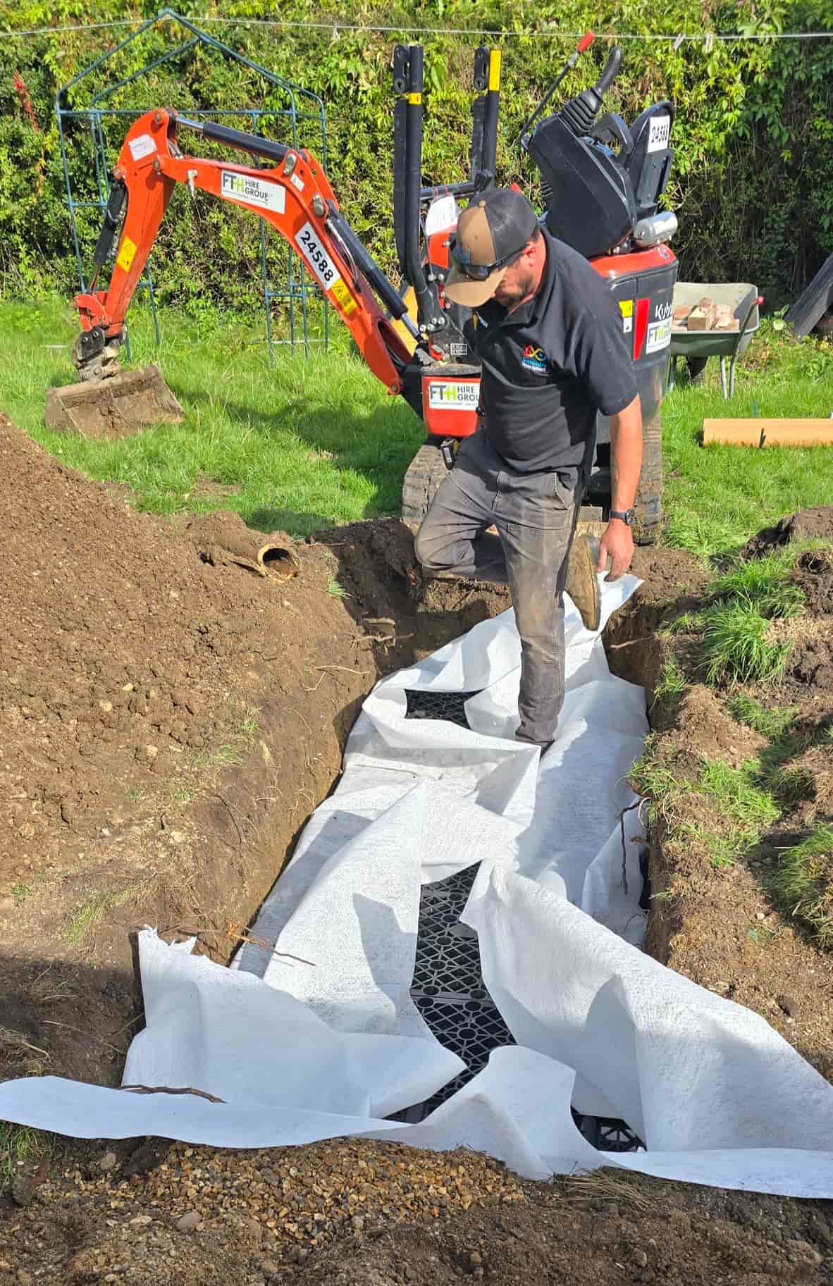 A man stands in a garden trench lined with white fabric and filled with drainage materials. A small orange excavator is parked nearby, and soil is piled on both sides of the trench.