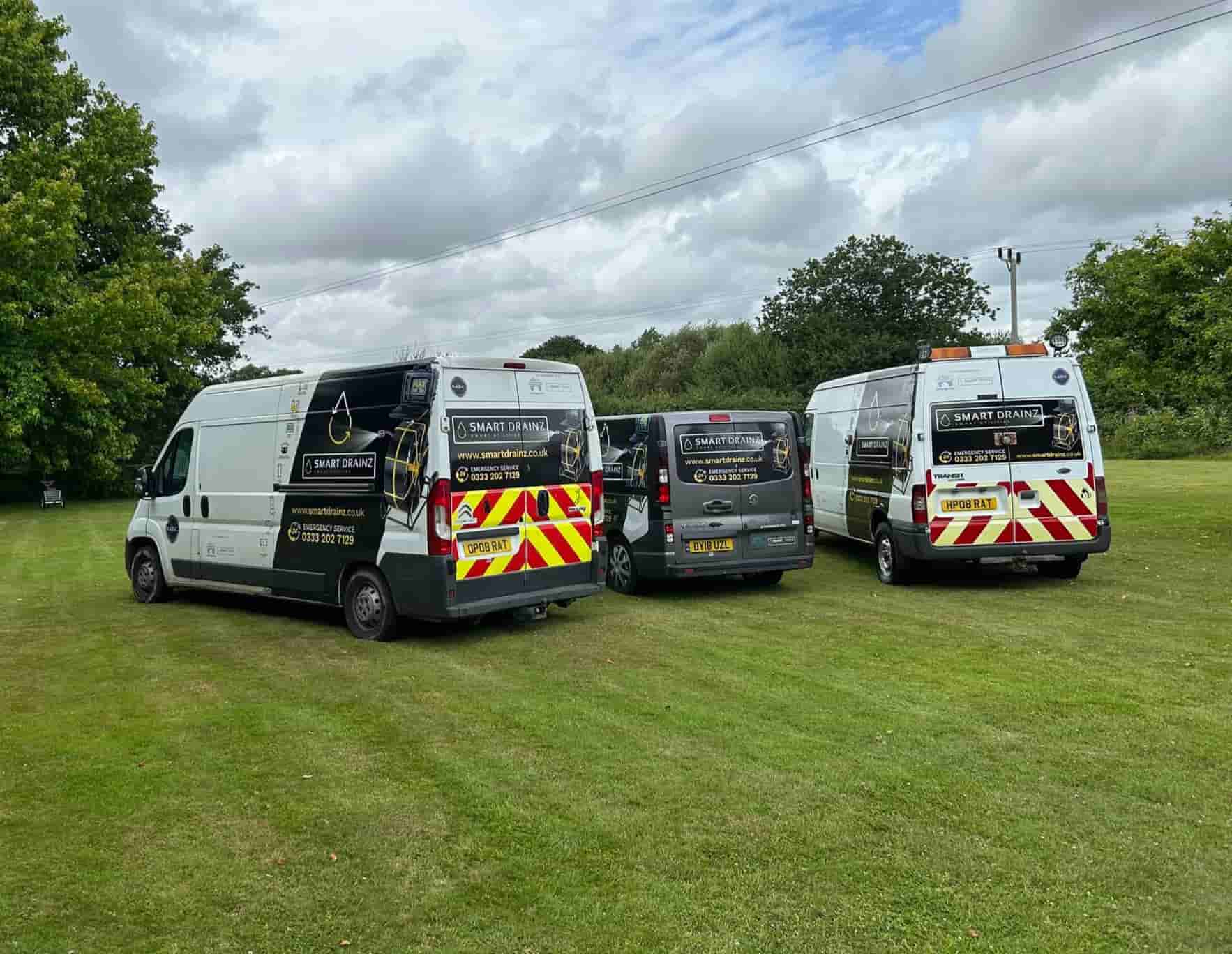 Three white work vans with company logos and high-visibility chevron patterns are parked on a grassy field under a cloudy sky, surrounded by green trees.
